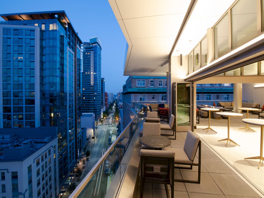 A modern rooftop terrace with tables and chairs has a railing overlooking a city skyline at dusk, featuring tall buildings and street lights.
