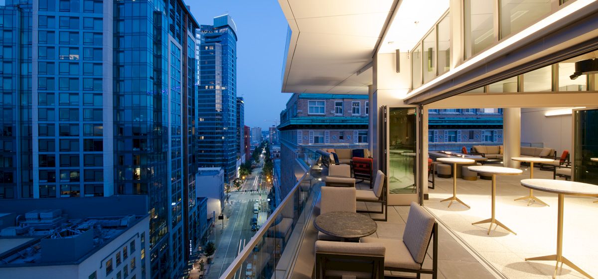 A modern rooftop terrace with tables and chairs has a railing overlooking a city skyline at dusk, featuring tall buildings and street lights.