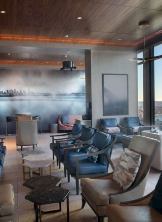 The image shows a modern lounge with comfortable seating, artistic decor on the wall, large windows offering a scenic view of a waterfront.