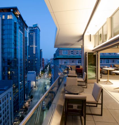 A modern cityscape at dusk featuring high-rise buildings and a contemporary balcony with seating areas and tables overlooking the urban scene.