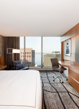 A modern hotel room with a large bed, wall-mounted TV, desk, chair, and expansive window offering a city and water view.