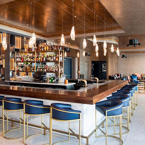 A stylish and modern bar with a long counter, blue bar stools, various bottles on shelves, pendant lights overhead, and a seating area in the background.