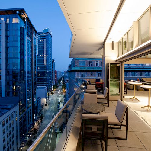 A modern balcony overlooking a cityscape at dusk, featuring tables, chairs, and open-air seating, with high-rise buildings in the background.