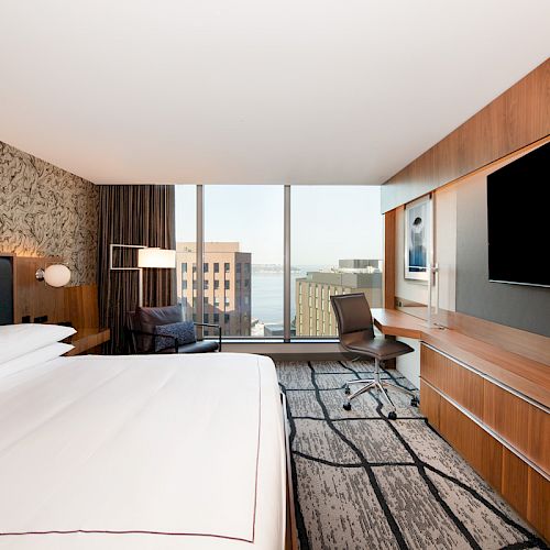 A modern hotel room features a large bed, wall-mounted TV, work desk, and chair, with a view of city buildings and the water from the window.