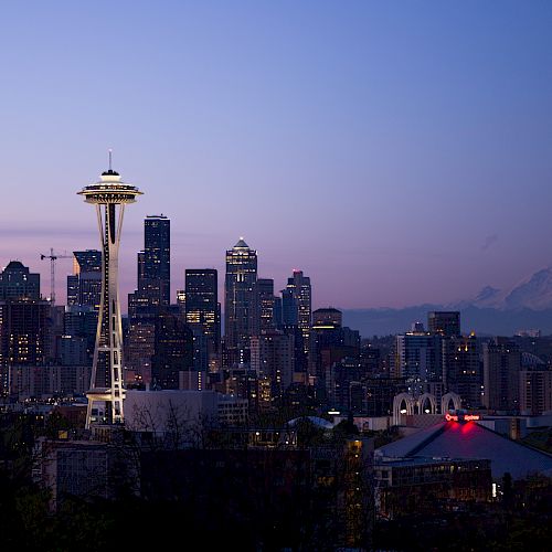 The image shows a skyline of a modern city at dusk, featuring a prominent observation tower, skyscrapers, and a distant mountain under a twilight sky.