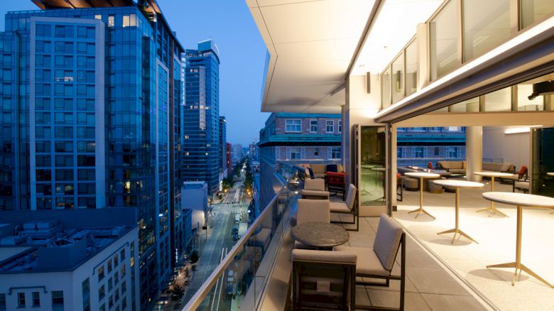 A rooftop terrace with seating overlooks a cityscape with tall buildings, lit street, and twilight sky. Modern and clean design.