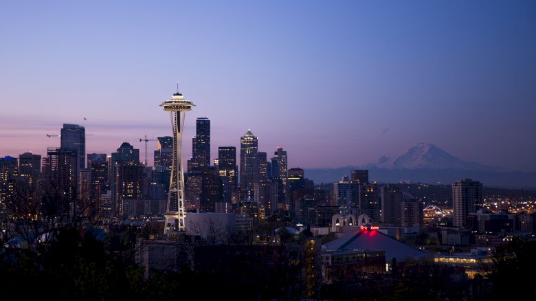 The image shows a city skyline at sunset, featuring the Space Needle, downtown buildings, and a distant mountain.