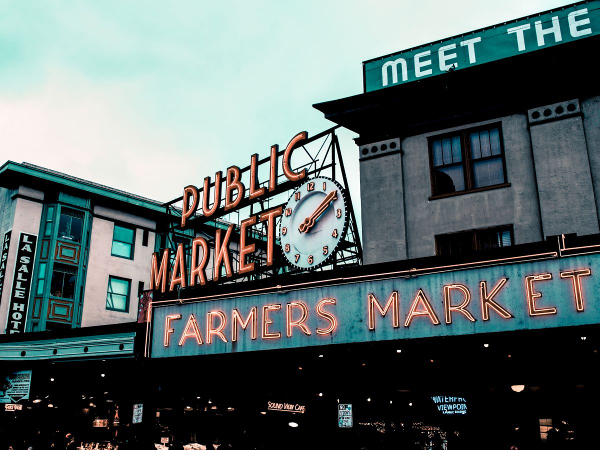 This image shows a public market sign with a clock and an illuminated "Farmers Market" sign in front of some buildings.