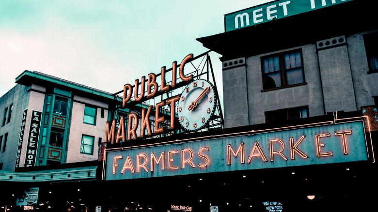 This image shows a public market sign with a clock and an illuminated "Farmers Market" sign in front of some buildings.