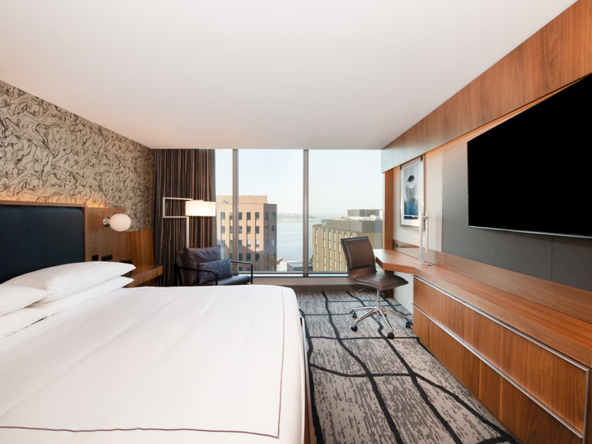 A modern hotel room with a large bed, desk, chair, wall-mounted TV, and a window offering a cityscape view and partial water view.