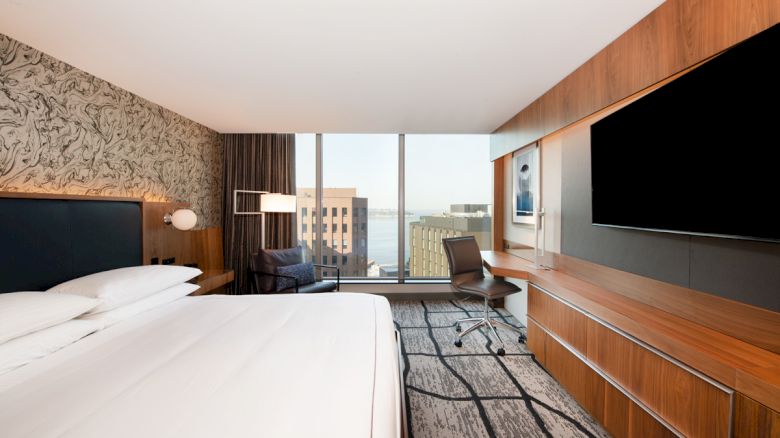 A modern hotel room with a large bed, desk, chair, wall-mounted TV, and a window offering a cityscape view and partial water view.