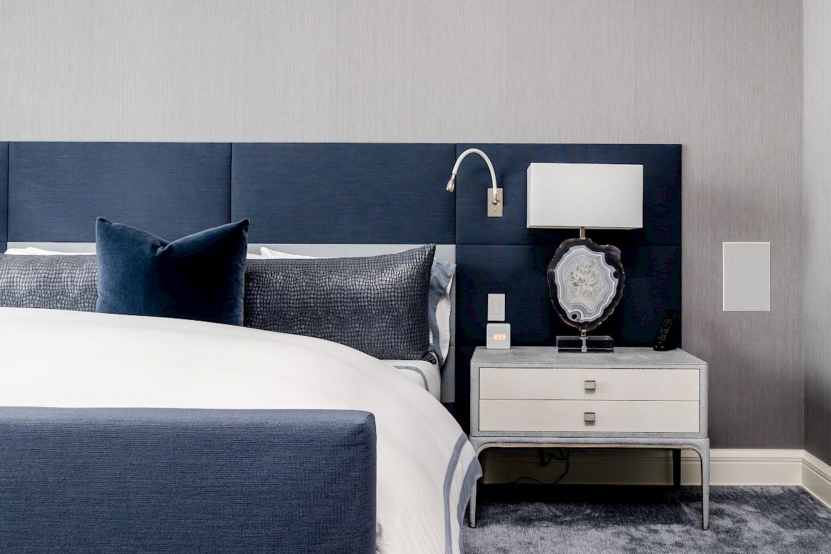 A modern bedroom features a blue upholstered headboard, white bedding, a nightstand with a lamp, and decorative items on a carpeted floor.