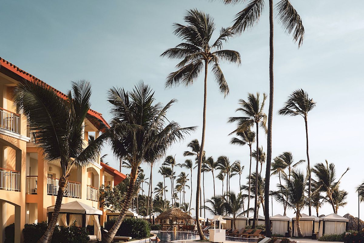 The image shows a tranquil resort scene with a pool, lounge chairs, tall palm trees, and a building with balconies under a clear, blue sky.