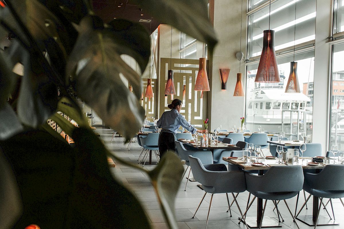 A person is arranging tables in a modern restaurant with stylish lighting, plants, and large windows overlooking a cityscape.