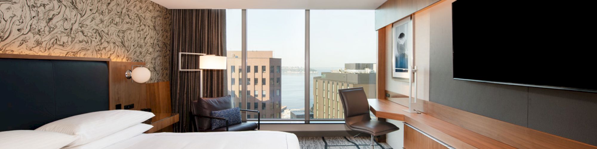 A modern hotel room with a large bed, desk, and chair, featuring a TV on the wall and a window with a city view.