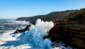 A dramatic coastal scene with large waves crashing against rocky cliffs, surrounded by a clear blue sky and dense forested hills in the background.