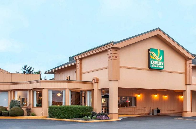 The image shows the exterior of a hotel building with a sign that reads "Quality Inn & Suites." The building has a pale color and is well-lit.