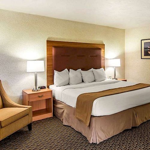 A hotel room with a large bed, four pillows, two nightstands with lamps, a brown chair, and a framed picture on the wall, ending the sentence.