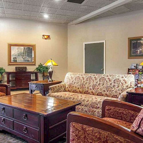 A cozy living room features patterned armchairs, a floral sofa, wooden coffee table, table lamps, framed artwork, and a small console table.