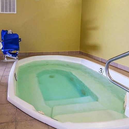 This image shows an indoor jacuzzi with a few steps leading in, a door, some chairs, and a blue lift chair for accessibility.