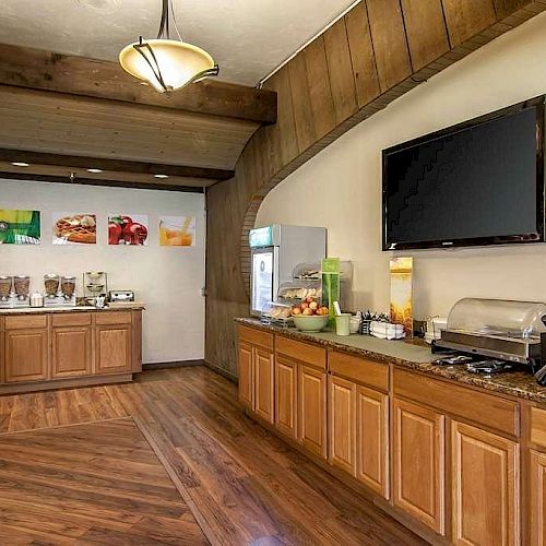 This image shows a breakfast buffet area with wooden cabinets, various food and drink dispensers, and a flat-screen TV on the wall.