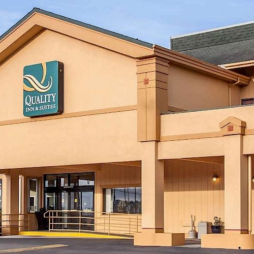 The image shows the exterior of a Quality Inn & Suites hotel. The building has a tan facade with a green sign and a covered entrance area.