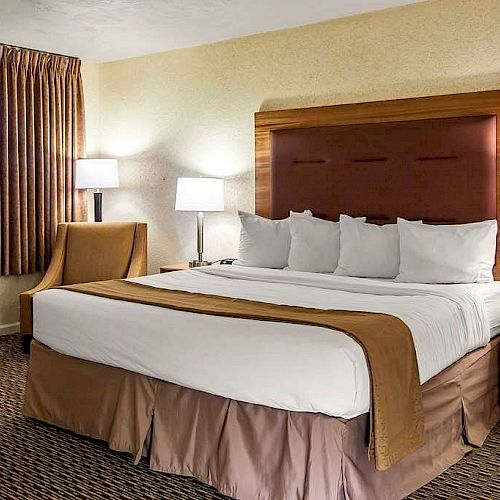 A cozy hotel room features a king-sized bed with white linens and brown accents, two bedside tables with lamps, and a window with curtains.