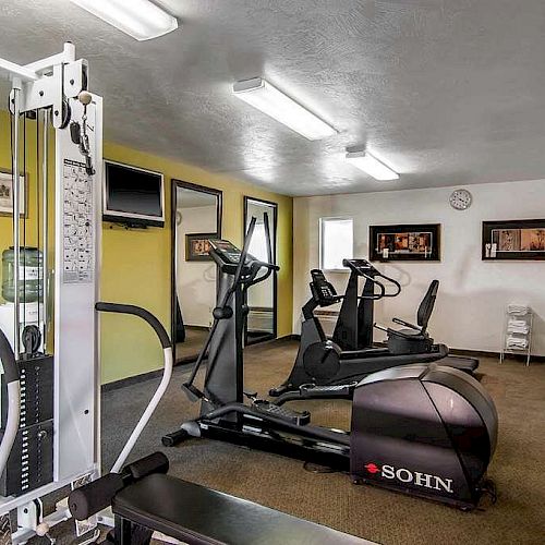 A small gym with exercise equipment including weight machines, stationary bikes, and a treadmill in a well-lit room with framed art on the walls.