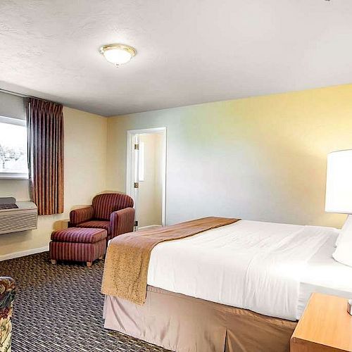 The image shows a hotel room with a bed, lamp, window, chairs, and an air conditioner, featuring simple decor and warm lighting.