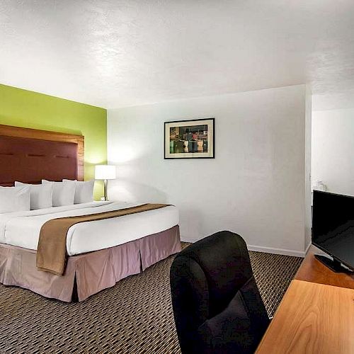 This image shows a tidy hotel room with a large bed, desk, TV, lamps, and a chair. The walls are painted green and white, with a framed picture.