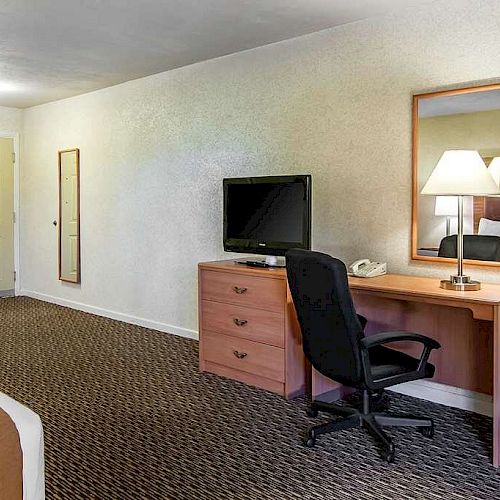 The image shows a hotel room with a desk, a TV, a chair, a lamp, a bed, a mirror, and a door, with carpeted flooring and beige walls.