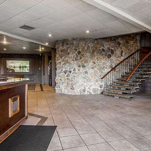 The image shows a spacious lobby with a reception desk, a stone wall, tiled floor, a staircase to the right, and a workspace with a chair.